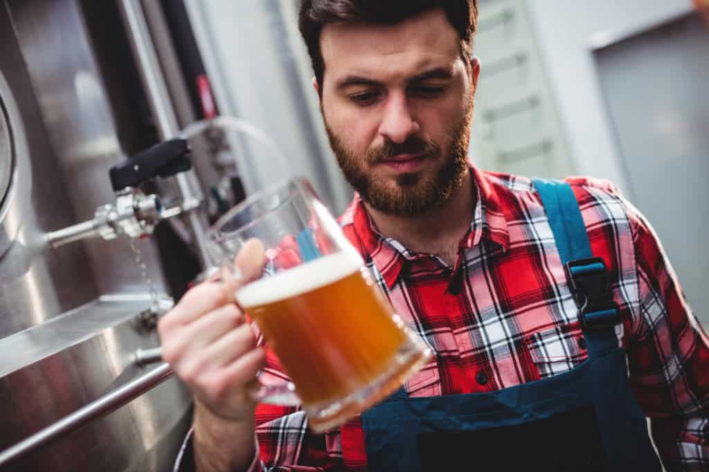 Manufacturer examining beer in glass by storage tank at brewery